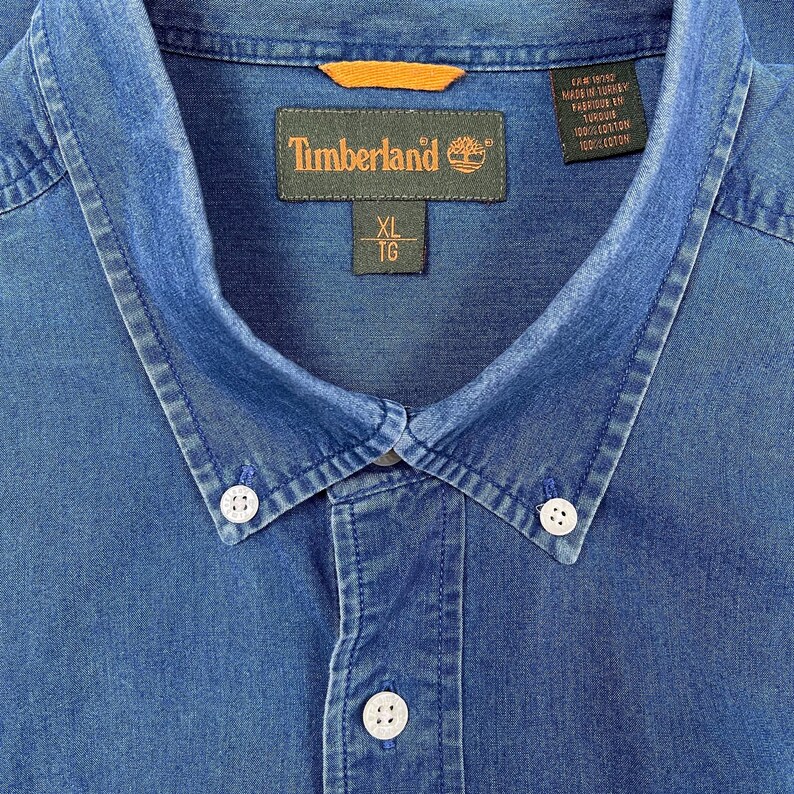 K&ouml;nnte beinhalten: Ein blaues Jeanshemd mit Knopfleiste und einem Timberland-Etikett. Das Etikett tr&auml;gt den Markennamen und die Gr&ouml;&szlig;e XL TG. Das Hemd ist in der T&uuml;rkei hergestellt und besteht aus 100% Baumwolle.