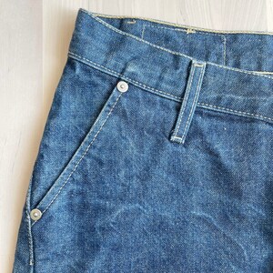May include: Close-up of the front of a pair of blue denim jeans. The jeans have a light wash and a classic five-pocket style. The waistband is visible, and the stitching is white.