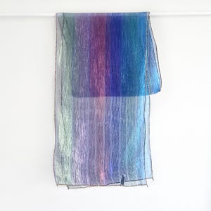 May include: A sheer, iridescent scarf with vertical stripes of color. The scarf transitions from green to purple to blue. The edges are finished with a black thread. The scarf is hanging against a white background.