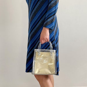 May include: A clear plastic tote bag with gold accents and the text "Tory Burch" printed on the front. The bag has two handles and is being held by a person wearing a blue and gray striped dress.