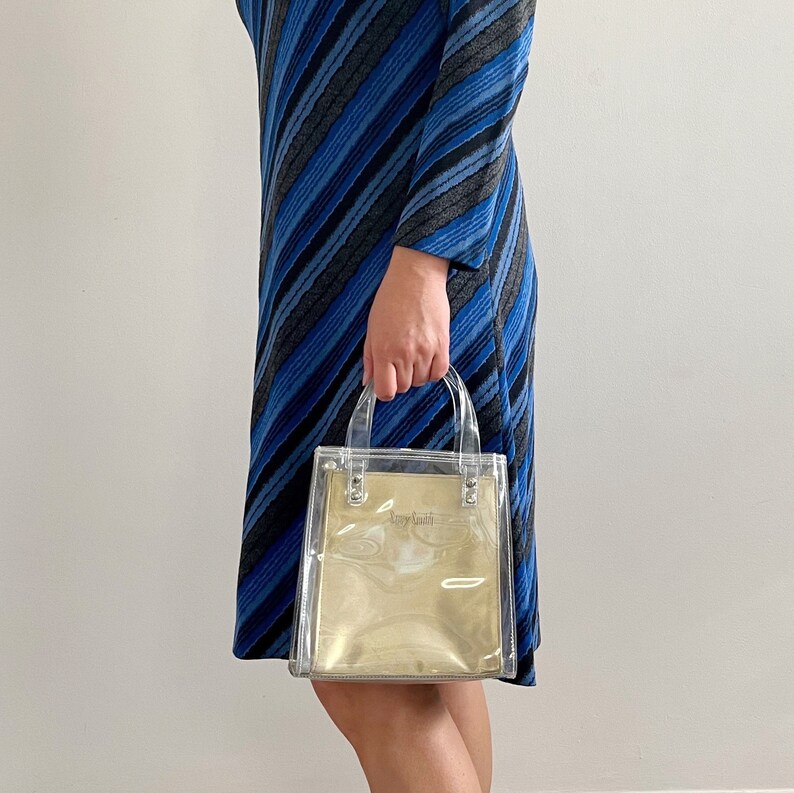 May include: A clear plastic tote bag with gold accents and the text "Tory Burch" printed on the front. The bag has two handles and is being held by a person wearing a blue and gray striped dress.