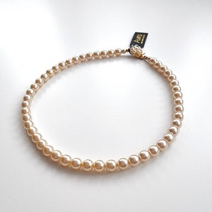 Puede incluir: Un collar de perlas de color crema con un cierre dorado. El cierre tiene un pequeño grupo de perlas falsas.