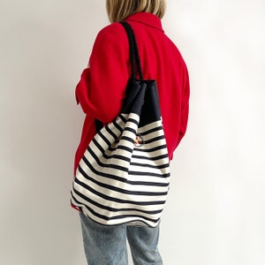 Puede incluir: Una bolsa de lona a rayas blancas y negras con un logotipo de estrella roja. La bolsa se lleva sobre el hombro de una persona que lleva una chaqueta roja.