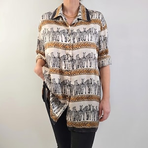 May include: A button-down shirt with a zebra print design. The shirt features a cream-colored background with rows of black and white zebras and brown decorative bands. The collar and cuffs have a dark brown trim.