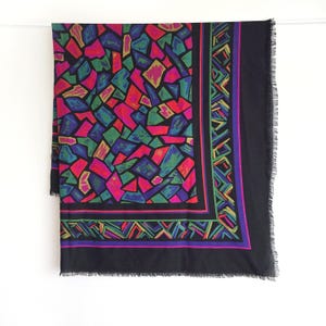 May include: A black scarf with a colorful geometric pattern. The scarf features a mosaic-like design in shades of red, green, blue, and yellow. The border has a geometric design with purple and green accents. The scarf has a fringed edge.