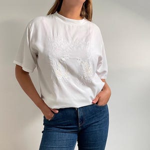 May include: White short-sleeved blouse with embroidered floral design and beaded accents. The round-neck blouse has a relaxed fit. A casual top suitable for everyday wear.