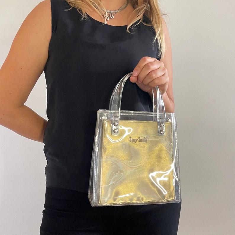 May include: A clear plastic tote bag with a gold lining and a silver logo that says "Tory Smith".