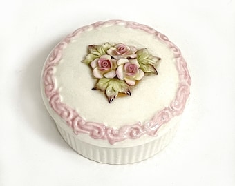 Puede incluir: Una caja de cerámica blanca con un diseño floral rosa. La tapa presenta un diseño en relieve de tres rosas rosas con hojas verdes.