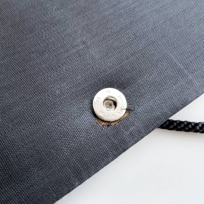 May include: Close-up of a black fabric bag with a silver snap closure. The snap has the text "K-SNAP" printed on it.