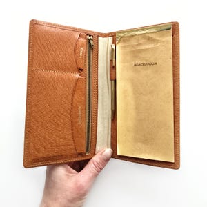 May include: Open, tan leather notebook with a pen, a notepad, and pockets for stamps and addresses. The notepad reads "MEMORANDUM". The notebook is open, revealing the interior. The leather has a textured appearance.