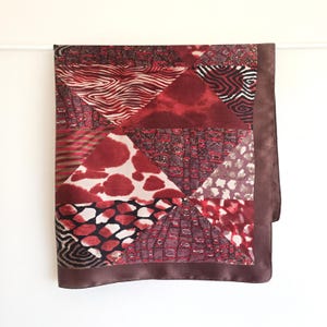 Op de afbeelding: Een vierkante sjaal met een patchworkontwerp van diverse dierenprints en geometrische patronen in tinten dieprood, bordeaux, zwart en crème. De sjaal is afgewerkt met een effen donkerbruine rand langs de zijkanten voor een duidelijk kader.