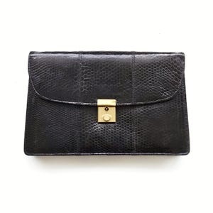 May include: A black clutch bag with a textured, reptile-like pattern. The rectangular bag has a curved flap and a gold-coloured clasp. The design includes vertical seams and a structured shape, suitable for carrying essentials.