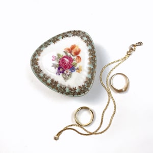 May include: A triangular porcelain trinket box with a white center featuring a colorful floral bouquet of pink, orange, and purple flowers, surrounded by a light green border with gold filigree, accompanied by a gold chain and two gold rings.