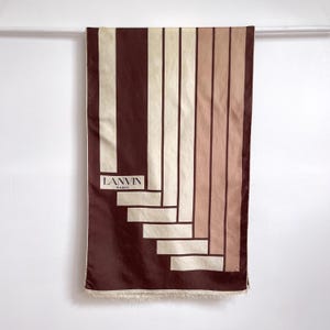 Könnte beinhalten: Ein rechteckiger Schal mit einem geometrischen Muster aus vertikalen und horizontalen Streifen in Dunkelbraun, Creme und Beige. Der Text LANVIN PARIS ist in einem schwarzen Rechteck auf der linken Seite gedruckt. Der Schal hat einen Fransenrand.