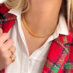 Op de afbeelding: Een gouden ketting met een delicaat, gevlochten ontwerp. De ketting wordt gedragen door een persoon met een wit shirt en een rood geruit jasje.
