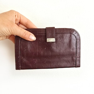 May include: A burgundy leather wallet with a rectangular shape and rounded corners. The wallet features a flap closure with a silver-colored metal clasp. The leather appears slightly worn, adding to its vintage appeal. The wallet is held against a white background.