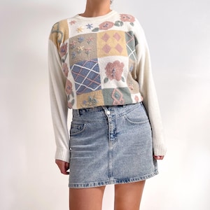 Puede incluir: Un suéter blanco con un diseño de patchwork con bordados florales en tonos rosa, azul y verde. El suéter tiene mangas largas y un cuello redondo.