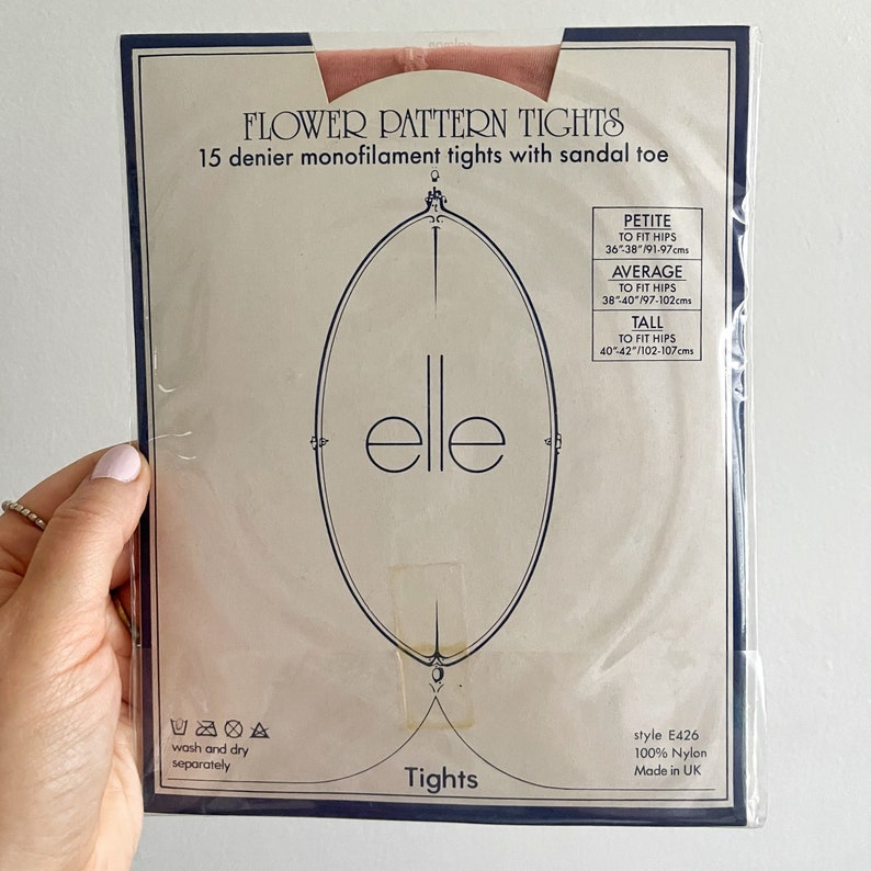 May include: Packaging for a pair of nude-colored tights with a floral pattern. The packaging is white with blue trim and features the word "elle" in a stylized font. The tights are 15 denier monofilament with a sandal toe. The packaging also includes size information for petite, average, and tall sizes. The text on the packaging reads "Flower Pattern Tights", "15 denier monofilament tights with sandal toe", "style E426", "100% Nylon", and "Made in UK".