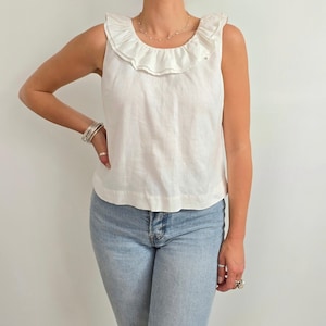 Puede incluir: Una blusa sin mangas de lino blanco con cuello con volantes. La blusa se combina con vaqueros de mezclilla azul claro. La modelo lleva pulseras y anillos de plata. La blusa es de diseño sencillo y elegante.