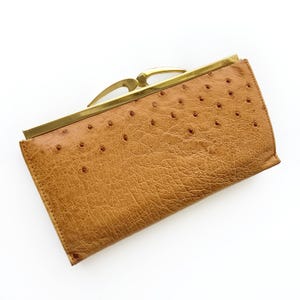 May include: A tan leather clutch bag with a textured surface and a gold-coloured clasp. The clutch has a rectangular shape with rounded corners and small, evenly spaced holes across the surface. The clasp is a curved design.