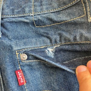 May include: Close-up of a blue denim pocket with a small red Levi's label and a metal button. The pocket has a small tear in the fabric.