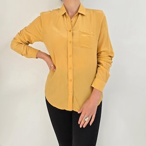 May include: Mustard yellow button-down shirt with a collared neckline and a single chest pocket. The long-sleeved shirt has buttoned cuffs and a curved hem. The shirt is paired with black pants.