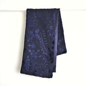 Puede incluir: Una bufanda azul marino con un estampado floral. La bufanda está hecha de un material suave y aterciopelado y presenta un diseño floral en relieve en un tono más oscuro de azul. La bufanda está drapeada sobre una superficie blanca.