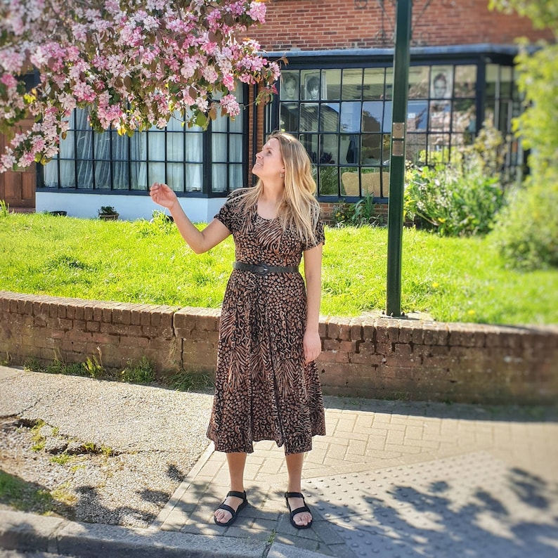 May include: A woman wearing a brown and black animal print midi dress with a black belt. She is standing on a sidewalk in front of a brick building with a large window.