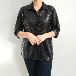 Puede incluir: Una camisa negra con botones y lentejuelas de manga larga. La camisa está hecha de una tela brillante y brillante.