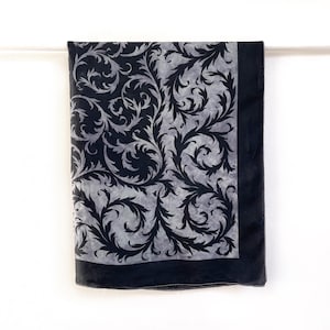 May include: A black and grey scarf with a detailed floral pattern. The scarf is rectangular with a dark border and a lighter grey background. The design features swirling leaves and vines.