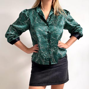 May include: Green and black striped button-down shirt with white accent lines. The shirt has a collared neckline, black cuffs, and puffed sleeves. The model is wearing a black denim skirt.