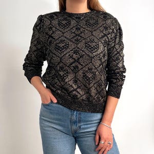 Può includere: Maglione nero e oro a fantasia geometrica, girocollo e maniche lunghe. Il maglione è realizzato in materiale lucido. La modella indossa jeans blu.