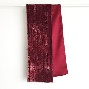 May include: A burgundy scarf with a textured, patterned design on one side and a smooth, solid burgundy fabric on the other. The scarf is hanging against a white background.
