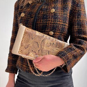 May include: A rectangular clutch bag with a snakeskin pattern in brown and gold tones. It features cream-coloured edges and a gold chain strap. The person is wearing a brown, black, and gold tweed jacket with gold buttons.