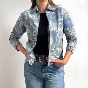 Könnte beinhalten: Hellblaue Jacke mit Reißverschluss und Landschaftsdruck. Die Jacke hat einen Kragen und lange Ärmel. Das Model trägt ein schwarzes Oberteil und blaue Jeans mit einem braunen Gürtel.