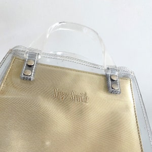 May include: A clear plastic tote bag with a beige fabric lining. The bag has a top handle and the brand name "Suzy Smith" is embroidered on the front.