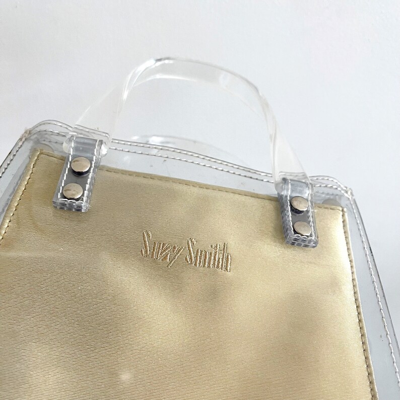 May include: A clear plastic tote bag with a beige fabric lining. The bag has a top handle and the brand name "Suzy Smith" is embroidered on the front.