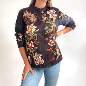 Könnte beinhalten: Dunkelbraune Jacke mit aufwendiger Blumenstickerei in Rot-, Orange- und Goldtönen. Die Jacke hat einen Stehkragen, lange Ärmel und dekorative goldene Knöpfe. Der Stoff hat ein dezentes geometrisches Muster.
