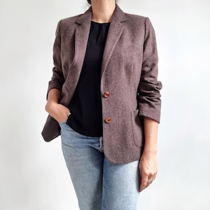 Puede incluir: Una chaqueta de tweed marrón con cierre de un solo botón y dos bolsillos delanteros. La chaqueta tiene un sutil patrón de espiga y se lleva sobre una camiseta negra y unos vaqueros azules.