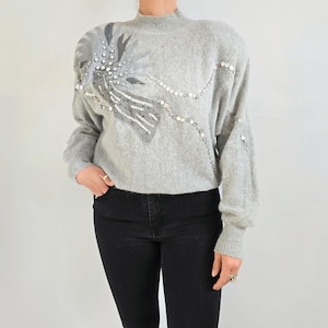 Può includere: Un maglione grigio chiaro con collo alto e maniche lunghe. Il maglione presenta un elaborato design sulla spalla sinistra, con paillettes e abbellimenti argentati. La modella indossa jeans neri.