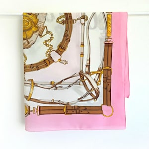 May include: A square silk scarf with a light pink border and a white center. The scarf features a detailed equestrian design with brown leather straps, gold accents, and a chain pattern.