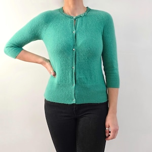 Puede incluir: Un cárdigan verde turquesa con mangas tres cuartos y cuello redondo. El suéter con botones tiene botones pequeños y redondos y un dobladillo acanalado. El cárdigan está hecho de un material suave y texturizado.