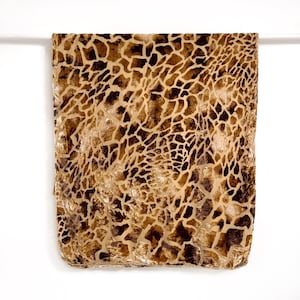 May include: A rectangular, folded fabric with a giraffe print pattern in shades of brown and tan. The fabric has a textured appearance and is set against a white background.