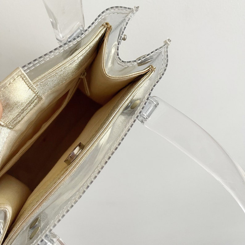 May include: A clear plastic tote bag with a gold lining. The bag has a top handle and a zipper closure.