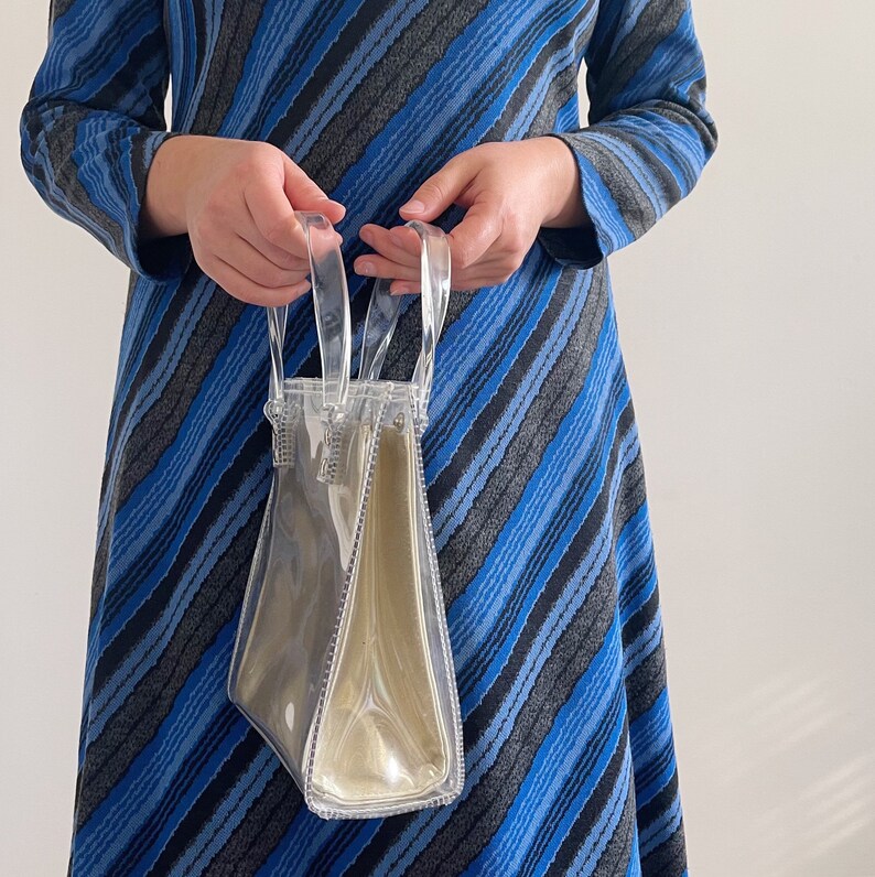 May include: A clear plastic tote bag with a zipper closure. The bag is being held by a person wearing a blue and gray striped dress.