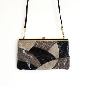 Puede incluir: Un clutch rectangular con un diseño de patchwork en tonos negros, grises y beige. El bolso tiene un marco dorado y una correa negra. El patrón de patchwork está hecho de varias formas y texturas.