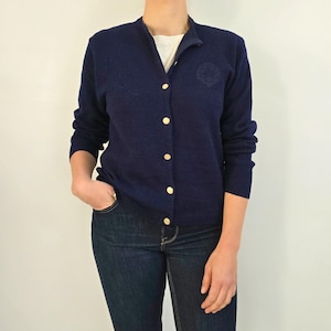 Può includere: Un cardigan blu navy con bottoni dorati. Il cardigan ha uno scollo tondo e maniche lunghe. Un piccolo stemma è visibile sul petto sinistro. La persona indossa jeans blu scuro e una maglietta bianca sotto.