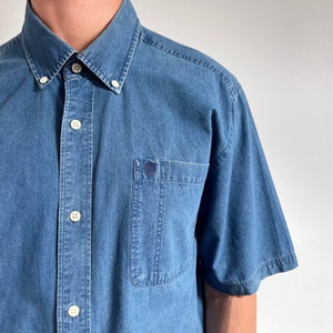 K&ouml;nnte beinhalten: Ein blaues, kurz&auml;rmeliges Hemd aus Denim mit einer Brusttasche und einem kleinen Logo auf der Tasche.