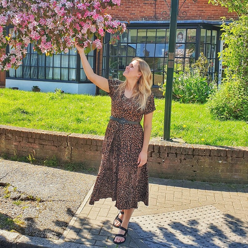 May include: A woman wearing a brown and black patterned midi dress with a black belt. She is standing on a sidewalk in front of a brick building with a flower bush.