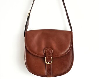 May include: A brown leather crossbody bag with a curved flap and a gold-coloured buckle. The bag has a long, adjustable strap and is suspended against a white background. The bag is a classic style.